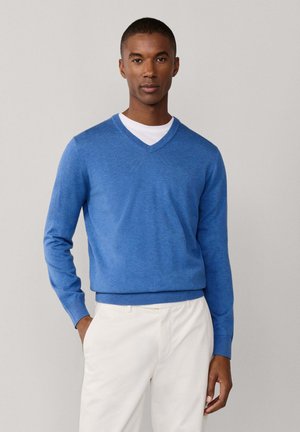 Hombre vestido con un suéter azul de cuello en V sobre una camisa blanca y pantalones blancos, de pie con una mano en el bolsillo frente a un fondo sencillo.