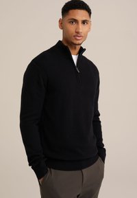 Svart zip-up-huvtröja i ett mjukt, strukturerat tyg. Har en hög krage och ribbade muddarna, ihop med byxor i neutrala färger.