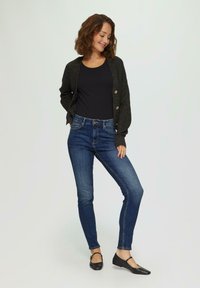 Donkergrijze cardigan met knopen over een zwart geribd topje, gecombineerd met blauwe skinny jeans en zwarte flats. Warme, gestructureerde breisels zijn te zien.