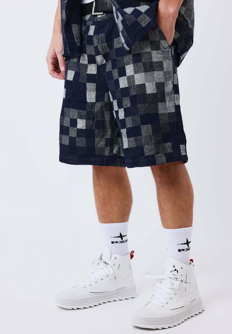 Homme portant un short à carreaux bleu marine et gris, des chaussettes blanches mi-mollet avec un logo noir, et des baskets montantes blanches à semelles texturées.