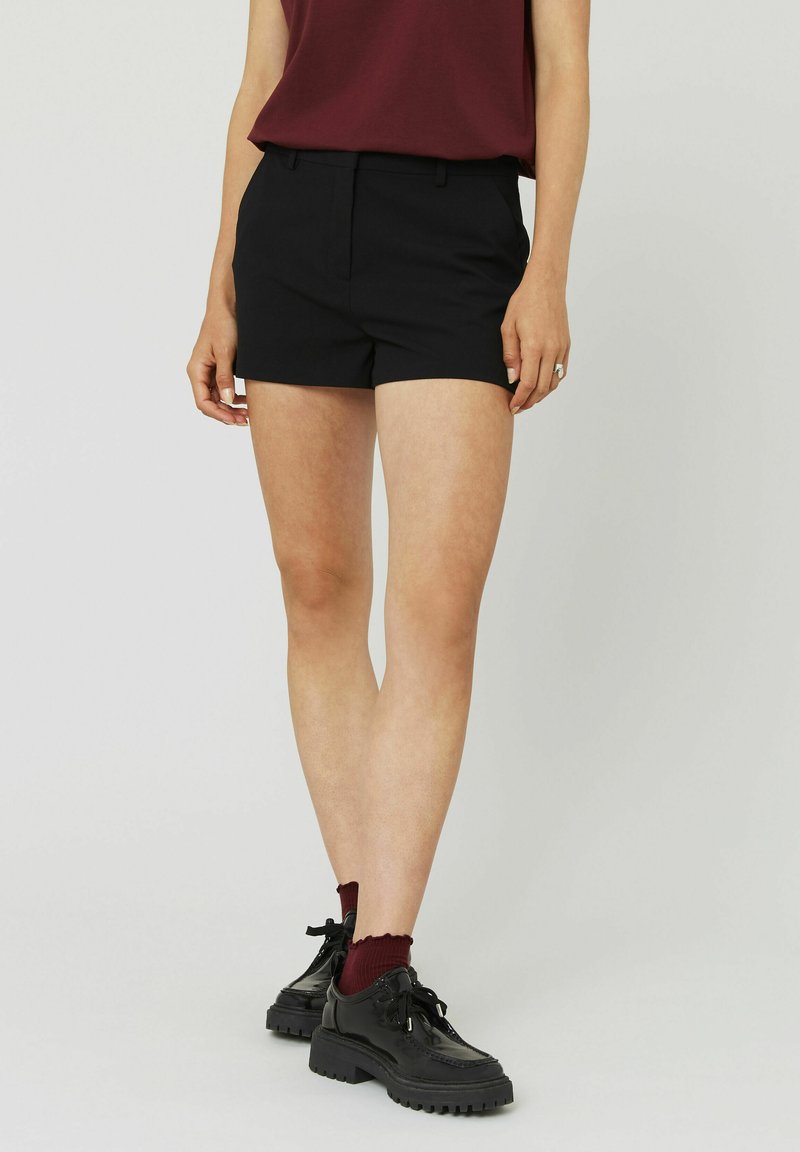 Shorts noirs ajustés avec une texture lisse, de longueur mi-cuisse, et des passants de ceinture. Associés à des chaussures noires à lacets et des chaussettes bordeaux.