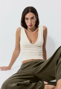 Femme aux cheveux foncés portant un haut blanc sans manches et un pantalon ample vert olive, assise devant un fond clair.