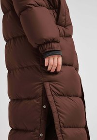 Bruine pufferjas met een lange silhoutte, elastische manchetten, zijopeningen en drukknoopdetails voor extra functionaliteit.