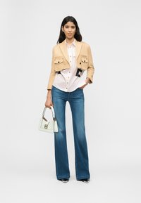 Γυναίκα που φορά cropped ταμπά σακάκι, λευκό πουκάμισο με κουμπιά, μπλε τζιν με καμπάνα, μαύρα και λευκά μυτερά παπούτσια, κρατώντας μικρή λευκή τσάντα.