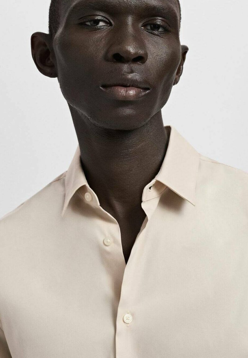 Chemise beige clair à boutons avec un col classique, texture lisse, et trois boutons visibles à l'avant, portée par une personne.