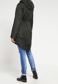 Dlouhý tmavě zelený nepromokavý kabát s kapucí, stažený v pase a s mírně delším zadním dílem. Nosit přes modré džíny a černé kotníkové boty.