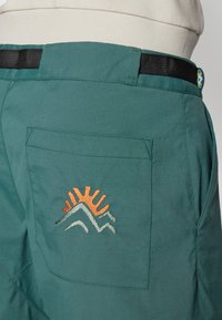 Zielone szorty cargo z tylną kieszenią, na której wyszyty jest zachód słońca i wzór gór w pomarańczowych, białych i zielonych nitkach. Czarny pas.