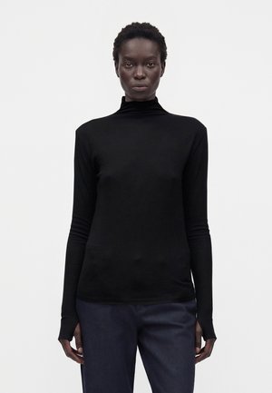 Femme aux cheveux courts et bouclés portant un col roulé noir à manches longues et un pantalon foncé, debout devant un fond clair uni.