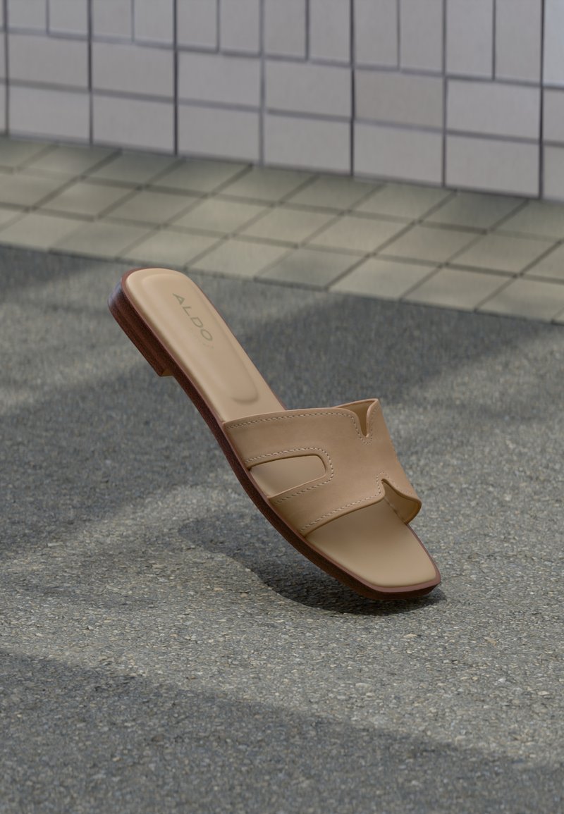 Beige skinnsandalett med utskjæringsdesign på strukturert fortausbakgrunn nær flislagt vegg.