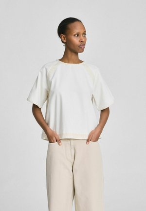 Femeie stând cu mâinile în buzunare, purtând un top alb larg cu mâneci scurte și pantaloni largi bej, privind într-o parte, pe un fundal simplu.