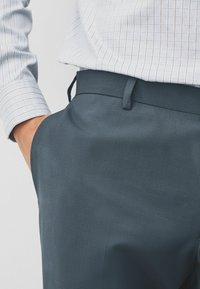 Dunkeltürkisfarbene Hosen mit glatter Textur, ausgestattet mit einem Bund mit Gürtelschlaufen und einer Hand in der Tasche. Ein leicht gemustertes Hemd ist sichtbar.