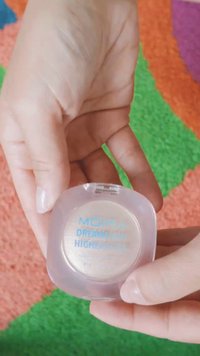 Hender som holder Moira Dreamlight highlighter-kompakt opp mot en fargerik teksturert bakgrunn.