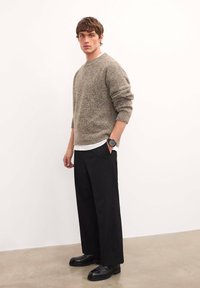 Pulover texturat gri cu mâneci lungi, asociat cu pantaloni negri largi și pantofi negri. Modelul stă în fața unui fundal alb.