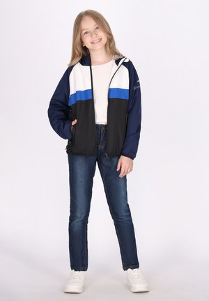 Giacca a vento nera e blu navy con strisce orizzontali bianche e blu; presenta un cappuccio e tasche laterali; abbinata a jeans blu e sneakers bianche.