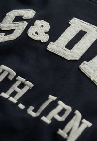 Marineblauwe stof met tekstuur, verhoogde witte letters die "S&D" en "NTH.JPN." lezen. Zichtbare stikdetails en lichte rafeling aan de randen.