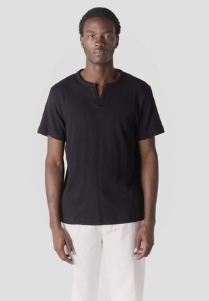 Uomo che indossa una camicia nera a maniche corte con scollo a V e pantaloni bianchi, in piedi contro uno sfondo chiaro e uniforme.