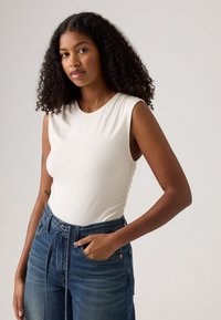 Top blanco sin mangas con escote redondo, combinado con unos vaqueros de denim azul de tiro alto que presentan un detalle de lazo en la cintura.