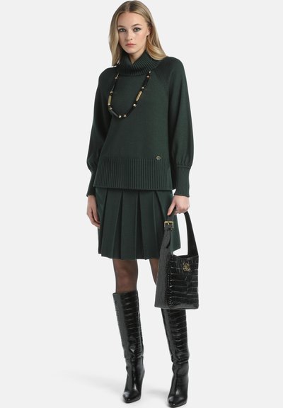 Pulover tricotat de culoare verde închis cu guler înalt, asortat cu o fustă plisată de aceeași culoare. Cizme negre până la genunchi și o geantă de umăr texturată neagră.