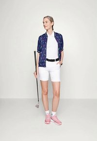 Sieviete, tērpusies baltos šortos un jūras zilā rakstu žaketē, tur golfa nūju, stāvot uz vienkrāsaina gaismas fona ar roku kabatā.