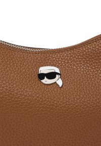 Borsa a tracolla in pelle marrone con finitura a ciottoli, dotata di una cerniera argentata e un pin in stile cartoon raffigurante una persona con capelli bianchi e occhiali da sole neri.