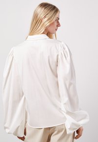 Camisa blanca de manga larga con mangas abullonadas y cuello clásico. Textura suave, ajuste relajado y dobladillo curvado. Diseño minimalista.