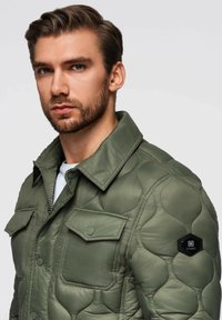 Uomo con capelli corti castani e barba, che indossa una giacca verde oliva trapuntata con tasche a soffietto e un patch esagonale nero sulla manica.