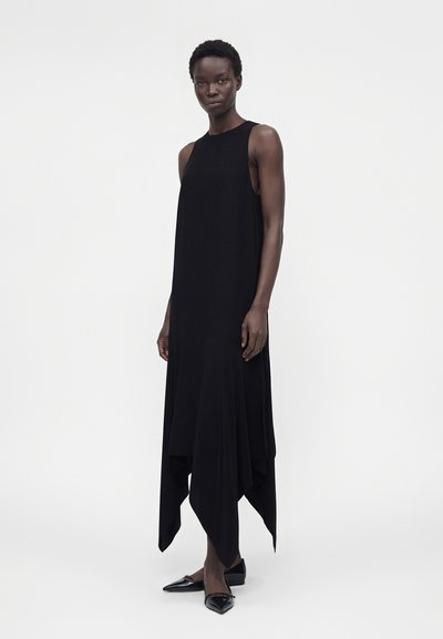 Γυναίκα που φοράει μακρύ μαύρο φόρεμα χωρίς μανίκια με ασύμμετρο τελείωμα και μαύρα μυτερά παπούτσια, στέκεται μπροστά σε λευκό φόντο.