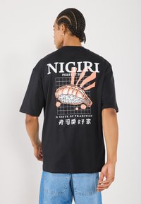 Fekete pamut póló nigiri sushi grafikai nyomattal, fehér és korall színű részletekkel, valamint szöveggel a hátsó oldalon. Rövid ujjú, laza szabású.