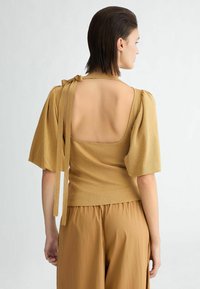 Top in maglia dorata con maniche a sbuffo, schiena scoperta e dettagli legati sulle spalle. Texture morbida, abbinato a pantaloni marroni larghi per creare contrasto.