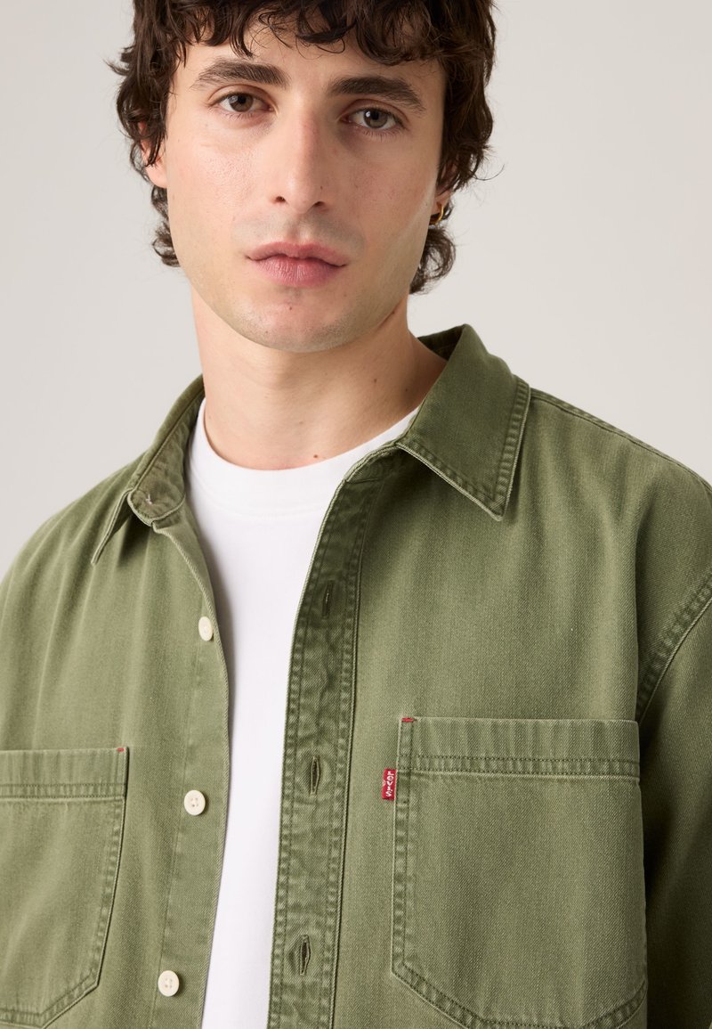 Giovane uomo con capelli ricci e scuri che indossa una camicia verde con bottoni sopra una t-shirt bianca su uno sfondo uniforme.