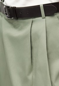 Κοντινό πλάνο ανοιχτοπράσινου πλισέ παντελονιού στερεωμένου με σκούρο καφέ δερμάτινη ζώνη με ασημένια πόρπη.