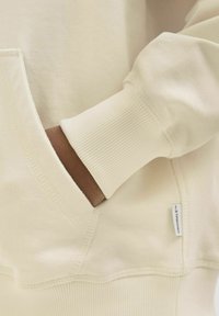 Krēmbalts sporta krekls ar ribotām aprocēm, priekšpusē ar kabatu un nelielu zīmola etiķeti, kas izšūta sānos. Gluda tekstūra.