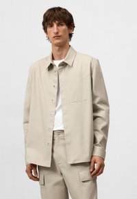 Camicia beige con bottoni realizzata in tessuto texture, dotata di colletto, maniche lunghe e taschino sul lato sinistro. Abbinata a pantaloni coordinati.