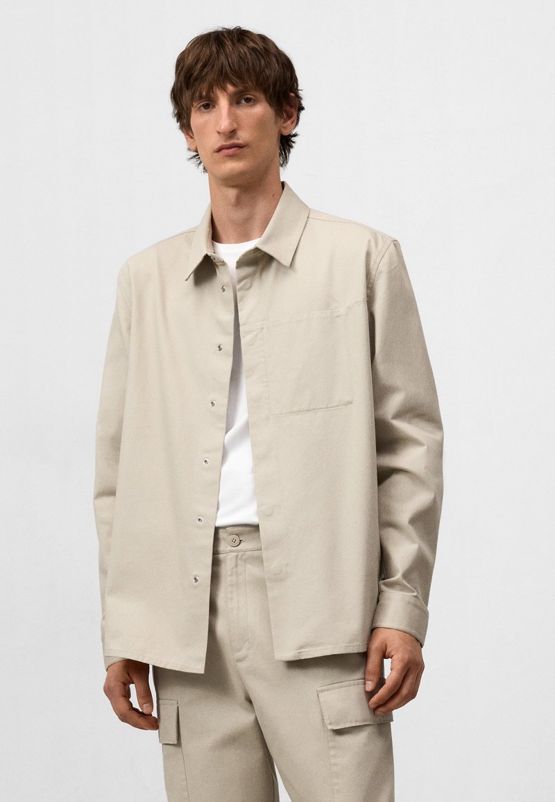 Camicia beige con bottoni realizzata in tessuto texture, dotata di colletto, maniche lunghe e taschino sul lato sinistro. Abbinata a pantaloni coordinati.