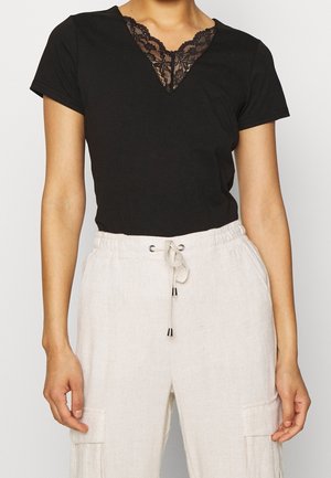 Czarna bluzka z krótkim rękawem z koronkowym dekoltem w kształcie litery V. Połączona z beżowymi, lnianymi spodniami z elastycznym pasem, które mają sznurki i boczne kieszenie.