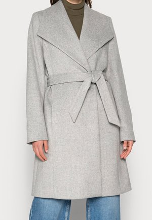 Femme portant un manteau en laine gris clair ceinturé par-dessus un col roulé olive et un jean bleu, debout les mains détendues le long du corps.