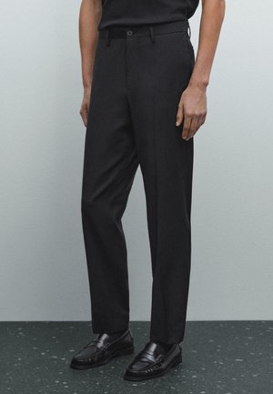 Osoba nosząca dopasowane czarne spodnie i czarne skórzane mokasyny, stojąca na jednolitym jasnoszarym tle.