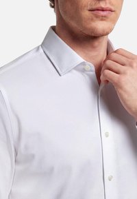 Mężczyzna poprawiający kołnierz białej koszuli, pokazujący zbliżenie szyi i górnej części tułowia na jednolitym tle.