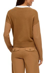 Brązowy sweter dzianinowy z ściągaczem u dołu i szerokimi rękawami. Biała kieszenna detal. Połączony z beżowymi spodniami z tylnymi kieszeniami zapinanymi na guziki.
