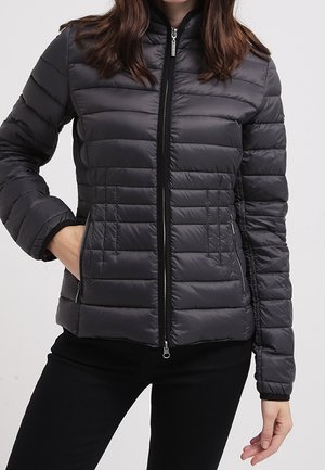 Kvinde iført en tætsiddende, sort, quiltet dunjakke med lynlås foran og lynlåslommer i siderne, den ene hånd i lommen, foran en ensfarvet baggrund.