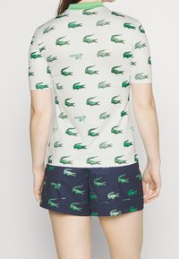 White polo shirt with green crocodile patterns, green collar, paired with navy shorts featuring similar patterns. Short sleeves, breathable fabric.