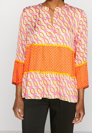 Kvinde iført en langærmet bluse med geometriske og polkaprikker i orange, pink og hvid, kombineret med sorte bukser.
