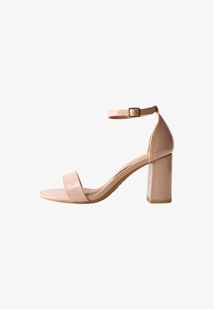 La sandale à talon en cuir verni nude présente un bout ouvert carré, une bride de cheville avec boucle et un talon épais pour plus de stabilité.
