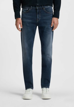 Dark blue denim jeans with a slim fit, featuring front pockets, subtle fading, and stitching accents. Paired with white sneakers.