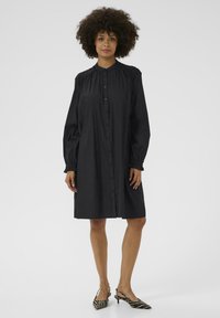 Robe noire à boutons avec manches longues, épaules smockées et coupe décontractée. Tissu texturé, longueur genoux, associée à des talons à imprimé zèbre.