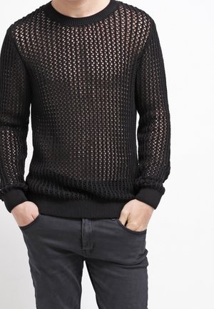 Mann trägt einen schwarzen, offen gestrickten Pullover über nackter Haut mit schwarzen Hosen, steht mit den Händen in den Taschen vor einem weißen Hintergrund.