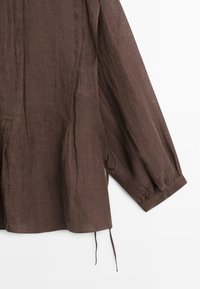 Bruine linnen blouse met lange mouwen, geplooide manchetten, verticale naaddetails en een koordje dat aan de zoom hangt op een witte achtergrond.