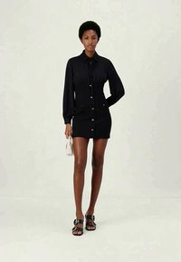 Abito nero a camicia con maniche lunghe, chiusura a bottoni frontale e colletto con coulisse. Indossato con sandali neri e una borsa a tracolla piccola. Texture liscia.