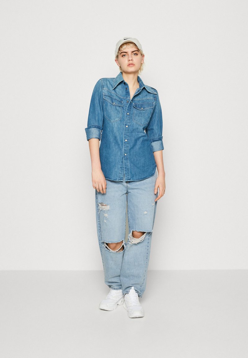 Denimhemd mit Kragen und zwei Brusttaschen, kombiniert mit hellblauen, ausgefransten Jeans mit großen Rissen. Weiße Sneakers vervollständigen das Outfit.