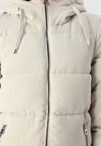 Beige, ribbet pufferjakke med hætte, der har en lynlås foran og sidelommer med lynlås. Den strukturerede stof giver et blødt udseende.